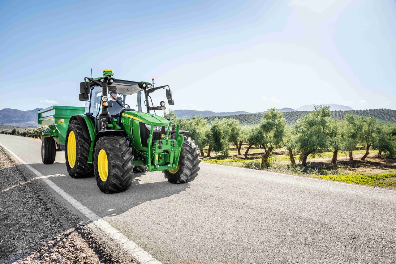 John Deere 5M med tilhenger på vei ved en olivenlund